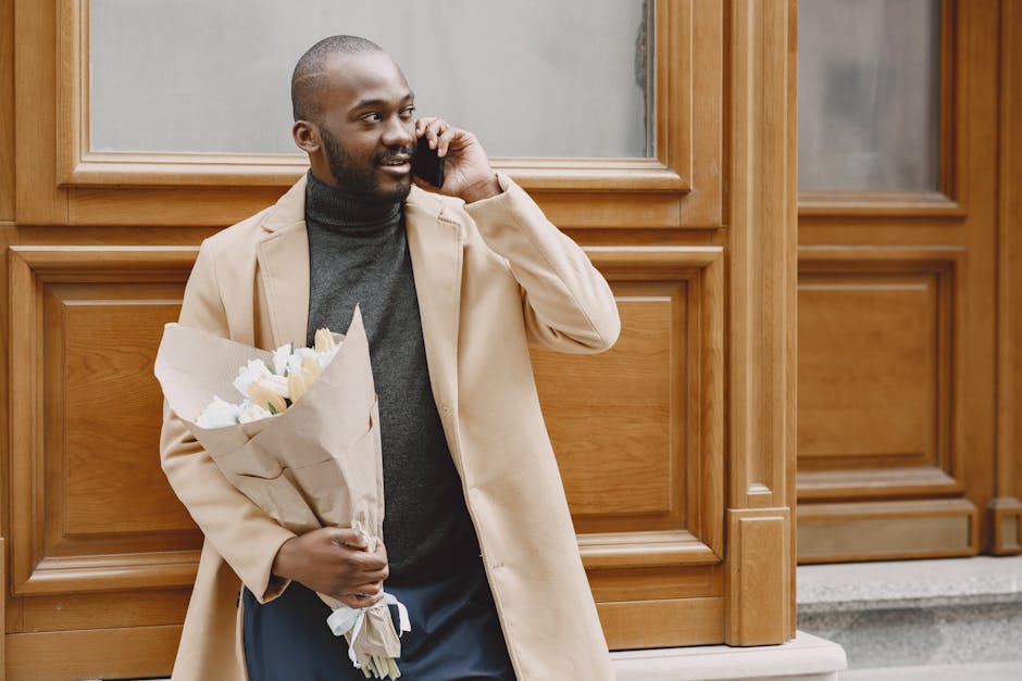 A man with dark skin, a beard, and a friendly expression is standing outside in front of a wooden panelled wall. He is holding a bouquet of fresh, cream and white flowers wrapped in brown kraft paper 