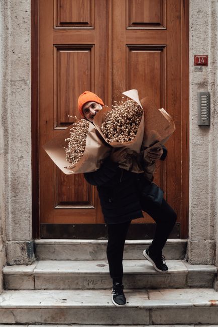 A person dressed in a black jacket, black pants, and black sneakers is standing on a marble doorstep in front of a large wooden door with a red number '14' plaque beside it. They are wearing an orange
