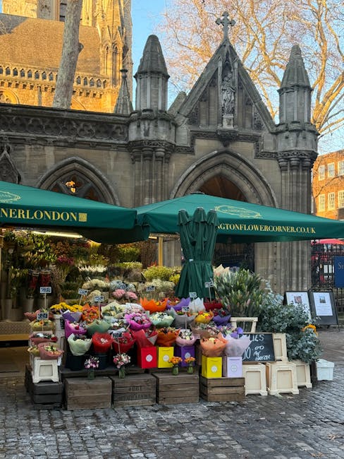 A vibrant flower stall situated outdoors in front of a historic church building with Gothic architectural elements, including pointed arches and decorative stonework. The stall is covered by green umb