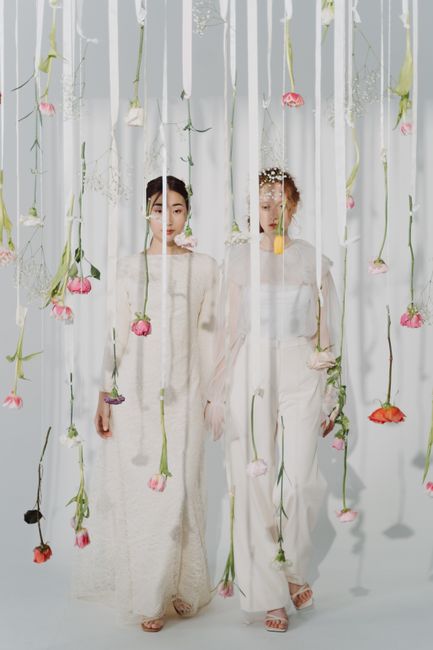 Two women dressed in elegant, light-colored dresses stand amidst hanging floral arrangements, with pink, peach, and white flowers suspended on green stems in a bright, softly lit environment. The flow