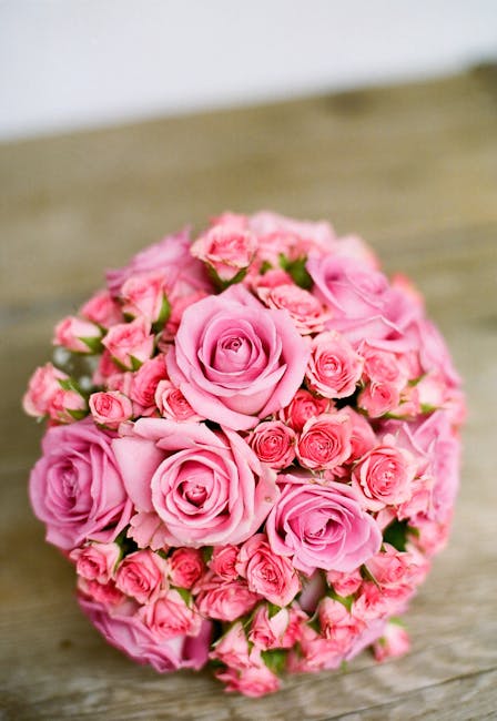 A round floral arrangement of fresh pink roses and smaller pink spray roses, densely packed with their petals slightly open to reveal the delicate interior. The bouquet is placed on a rustic wooden su