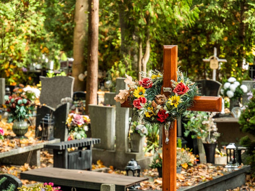 A wooden cross decorated with a vibrant floral arrangement, featuring red roses, yellow daisies, and pale pink flowers, is situated in a cemetery with various gravestones and memorial plaques. The flo