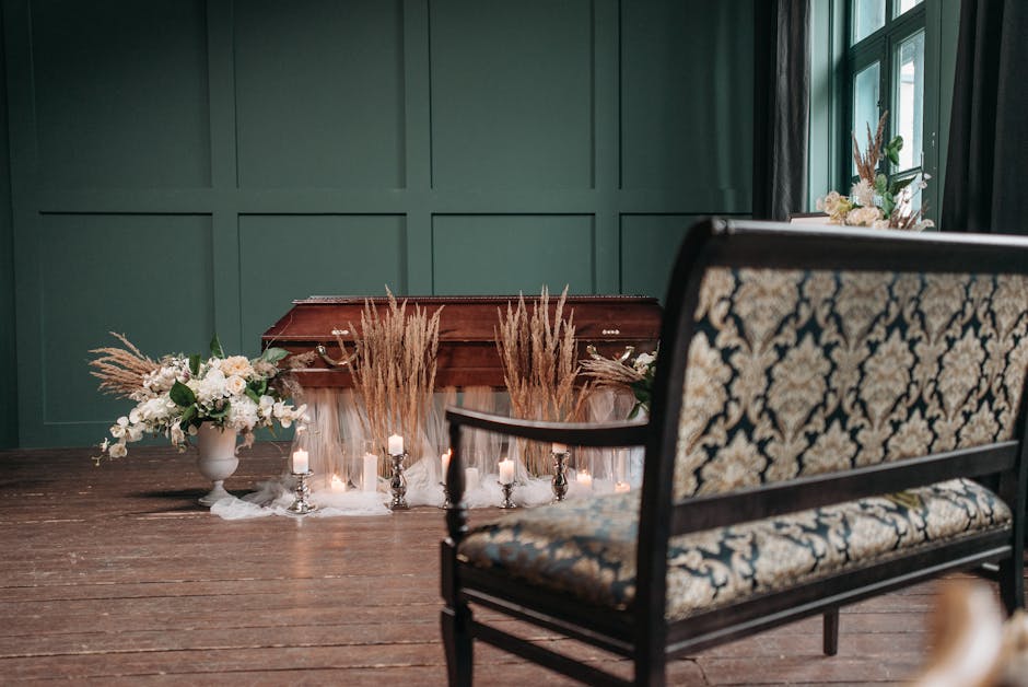 A floral arrangement with white roses, lilies, and greenery in a white pedestal vase, complemented by dried pampas grass in a larger white urn, positioned on a wooden floor near a green paneled wall a